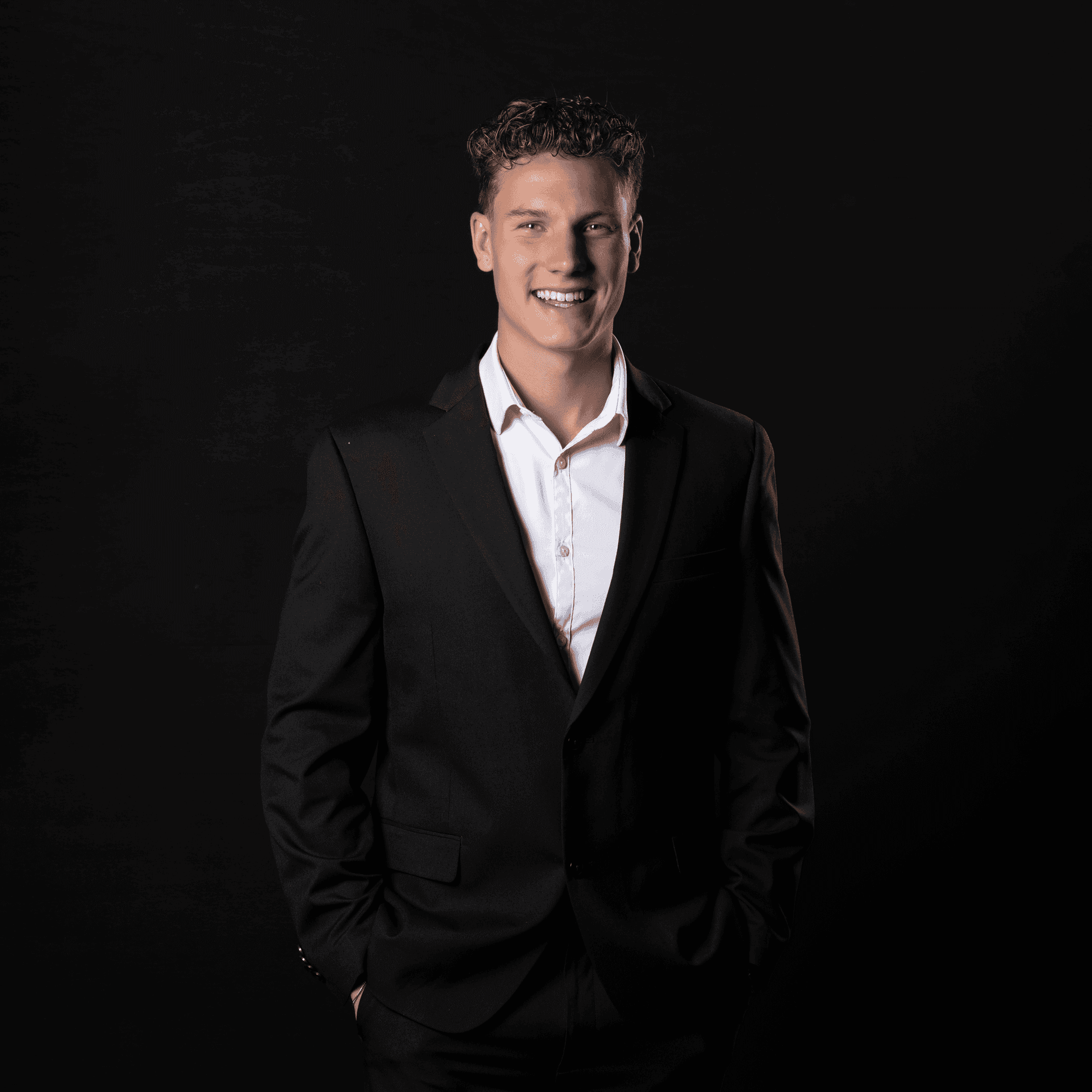 Smiling person in a black suit and white shirt against a dark background.